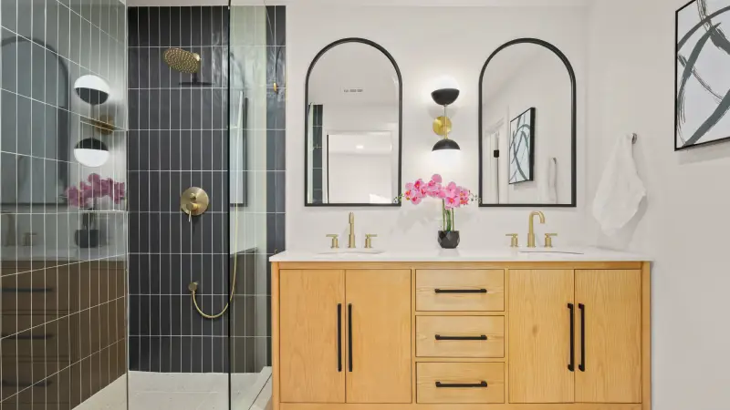 After image of a remodeled restroom where the shower is tiled has glass doors, bronze handles and shower head. The bathroom also two mirros with the top corners bering rounded and a vanity with his and her sinks and drawers.