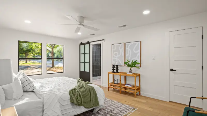 A remodeled master bedroom featuring a bed on a rug, the room has in celing lights as well as a celing fan the restroom has a sliding barn door style door. Two pattern paintings hang across from the bed.